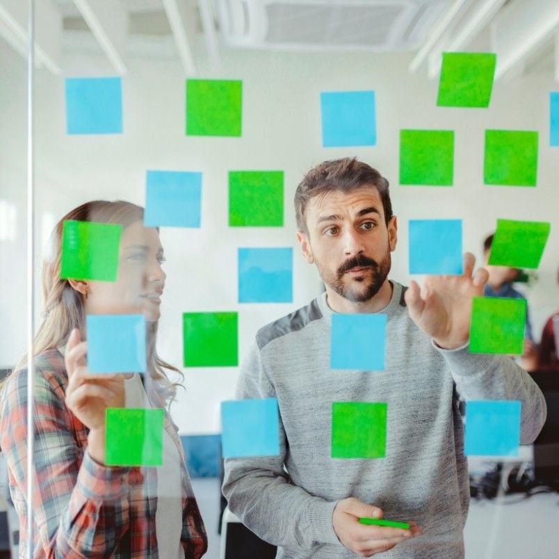 Two people stand behind a glass wall covered with blue and green sticky notes, demonstrating agile skills as they discuss and point at the notes while collaborating in a bright, modern office environment.