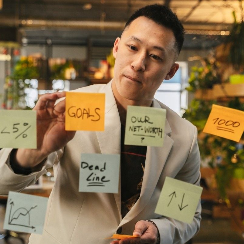 A man in a white blazer demonstrates agile skills as he writes on glass covered with colorful sticky notes labeled “GOALS,” “OUR NET-WORTH,” and more, surrounded by charts, symbols, and plants in a modern office.