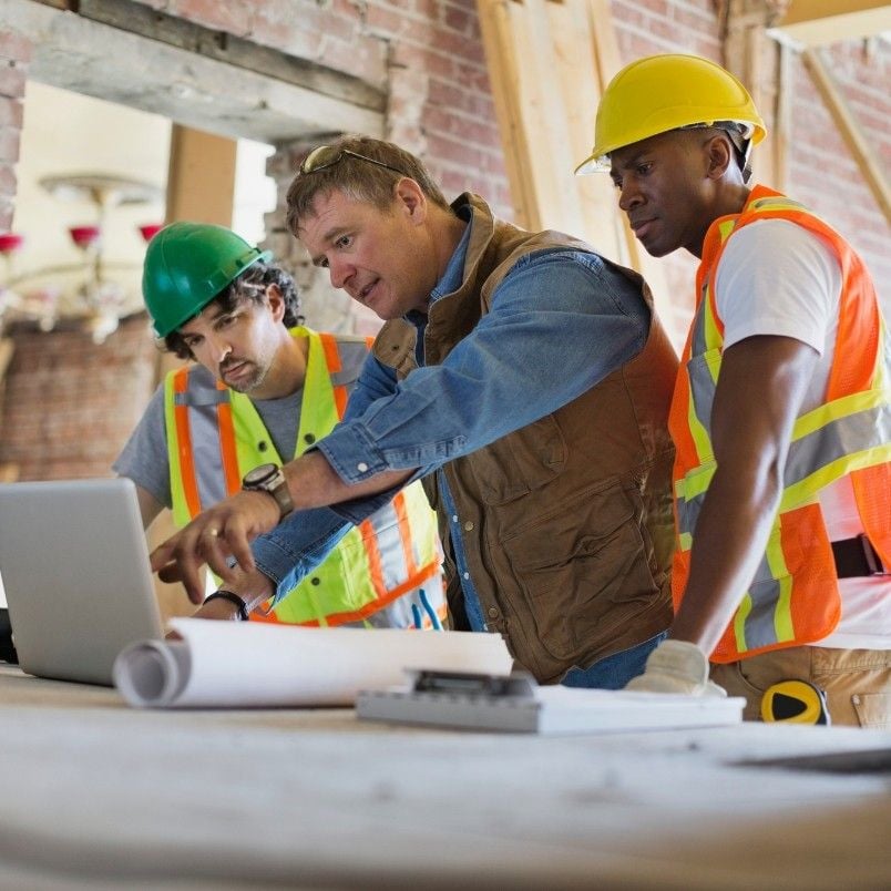 Three construction workers wearing safety vests and hard hats review building plans and look at a laptop, demonstrating effective Construction Project Management on a work site with exposed brick walls and wooden beams.