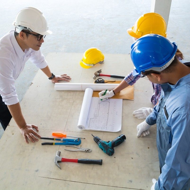 Three people in hard hats and safety gear review blueprints on a table with construction tools, showcasing effective Construction Project Management. A yellow hard hat rests nearby as they discuss plans and strategies.