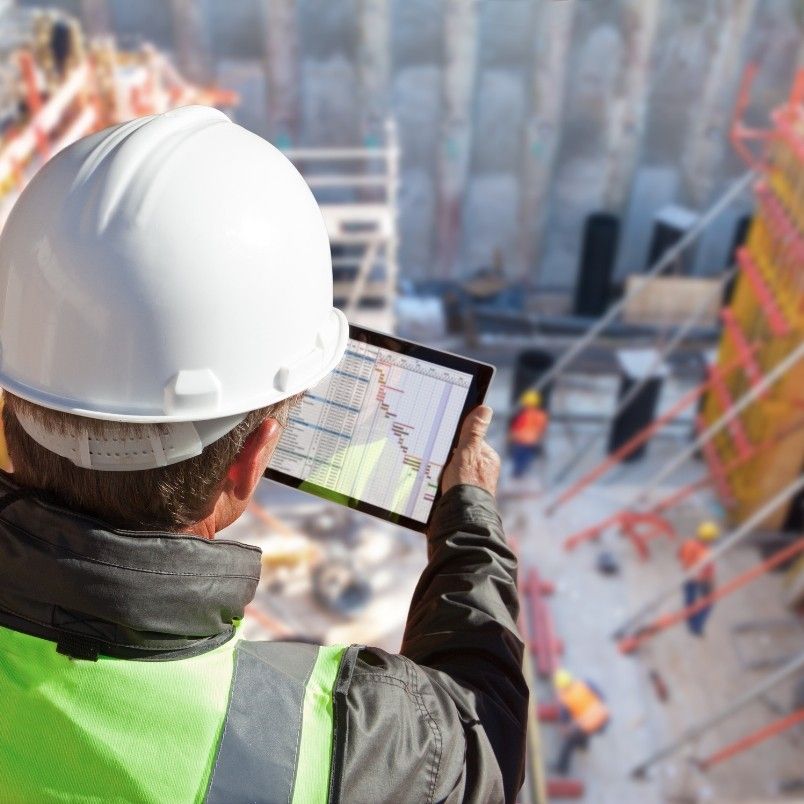 A person in a safety vest and hard hat checks a tablet displaying a Construction Project Management plan at a busy construction site, with workers and materials visible in the background.