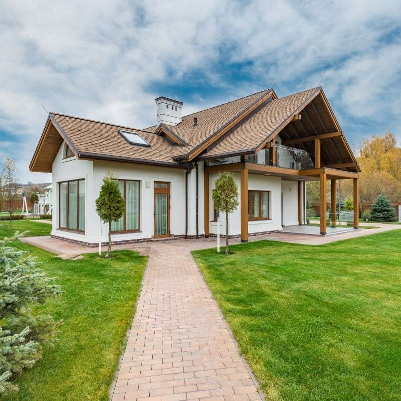 Modern two-story house with large windows, wooden pillars, and a covered balcony—an impressive result of expert Construction Project Management—surrounded by a well-kept green lawn, trees, and a paved walkway under a partly cloudy sky.