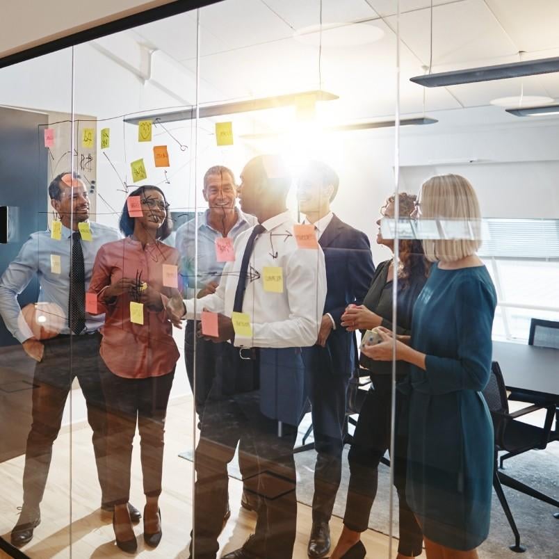 A group of six business professionals with agile skills stand in a glass-walled office, smiling and discussing ideas while placing colorful sticky notes on the glass. Sunlight shines through the window, creating a bright, collaborative atmosphere.