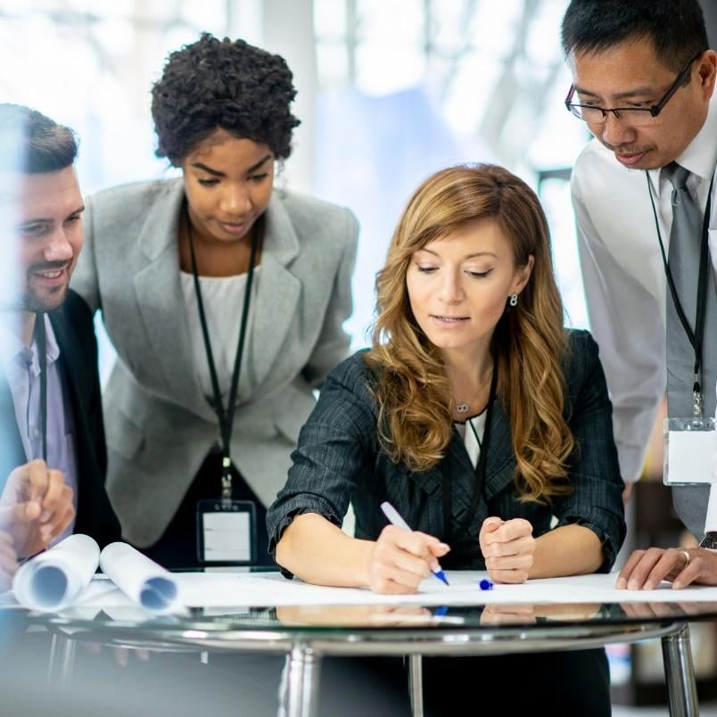 Four business professionals, three standing and one seated, collaborate around a table with documents. The seated woman writes as the others discuss the project charter in an agile setting, all appearing engaged and focused.