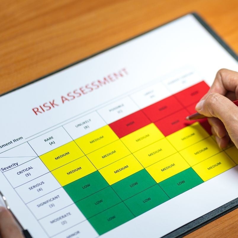 A hand holds a pen over a risk assessment chart on a clipboard, displaying a color-coded matrix and referencing the risk register to identify varying risk levels of different severity and likelihoods.