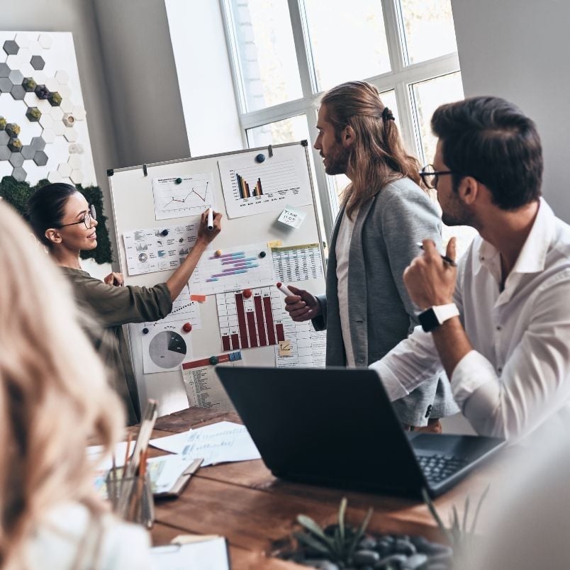 Four people are in a modern office. One woman points at charts and graphs on a whiteboard, presenting data from an Agile Project Charter Template to colleagues. Two men listen attentively, one with a laptop as sunlight streams through large windows.