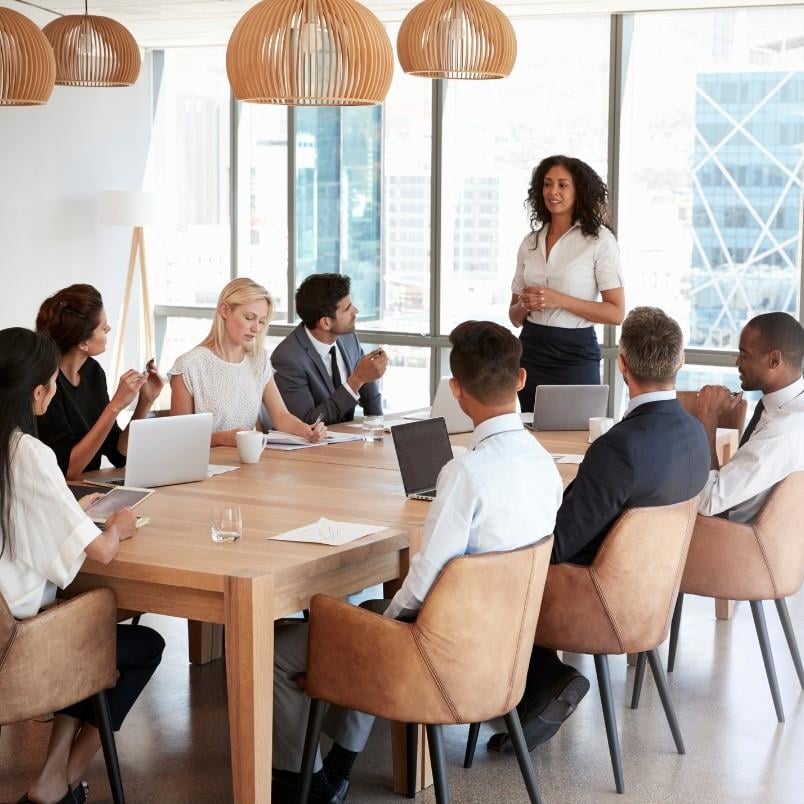 A woman stands and speaks to eight colleagues seated around a conference table in a modern office with large windows and natural light, as they collaborate on their Agile Team Charter. Laptops and documents are spread across the table.