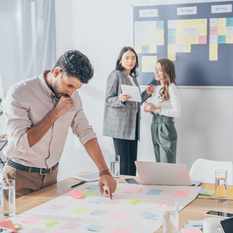 A man stands at a table covered with colorful sticky notes, deep in thought, embodying the project manager role in Scrum, while two women in the background discuss papers near a board with organized sticky notes and task columns.