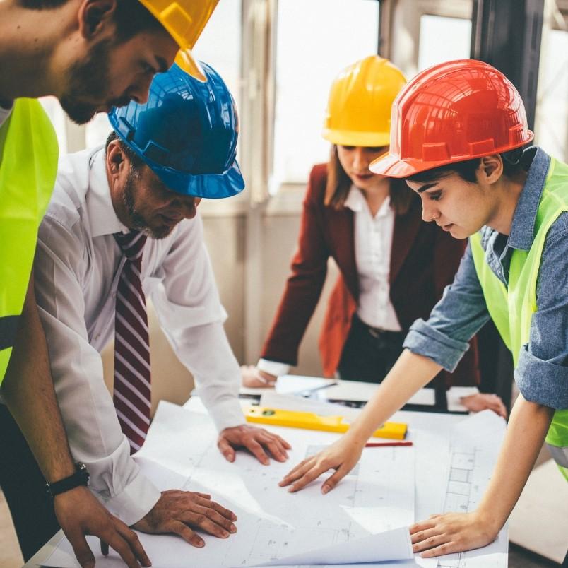 Four people, wearing safety helmets and vests, gather around a table studying blueprints and construction project management plans in a brightly lit room. Various construction tools are also placed on the table.