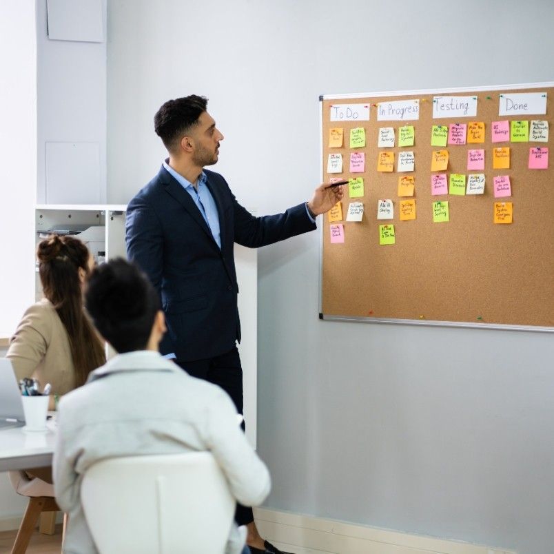 A man in a suit points to a corkboard with colorful sticky notes labeled "To Do," "In Progress," "Testing," and "Done" as he discusses the Agile Team Charter while two people seated at a table listen attentively.
