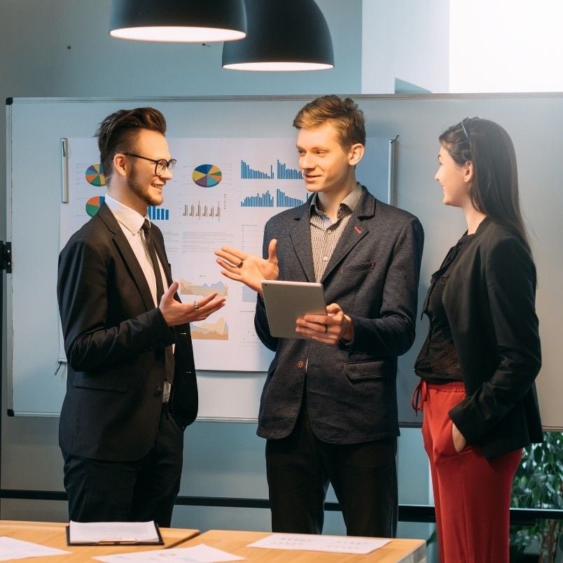 Three young professionals stand and talk in an office setting, with one holding a tablet. Behind them, a whiteboard displays colorful charts and graphs, reflecting their agile skills as they engage in a productive and upbeat discussion.
