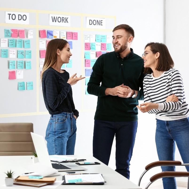 Three people stand and talk in front of a Kanban board with sticky notes labeled "To Do," "Work," and "Done"—an agile project management example. A laptop and notebooks are on the table beside them in the office.