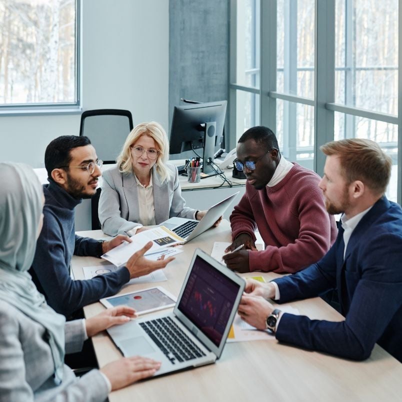 Five people in business attire sit around a table with laptops and documents, reviewing an Agile Project Charter Template in a bright, modern office with large windows.