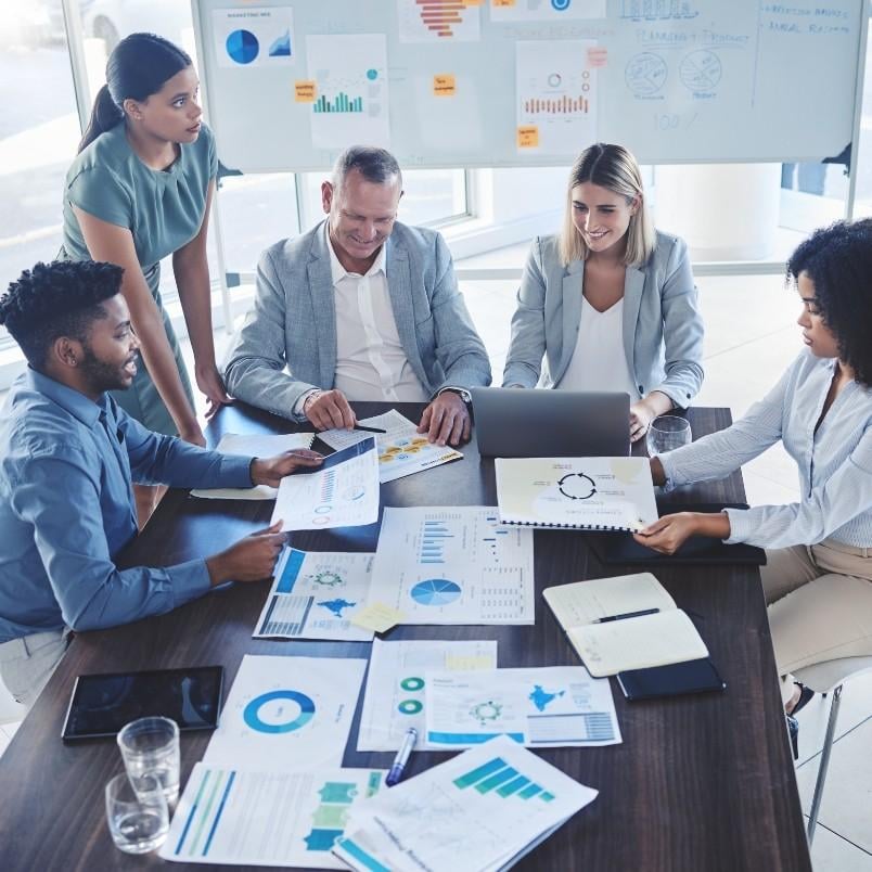 Five professionals gather around a table covered with charts, graphs, and documents, discussing how to track project progress. A whiteboard with graphs is visible in the background as one person types on a laptop.