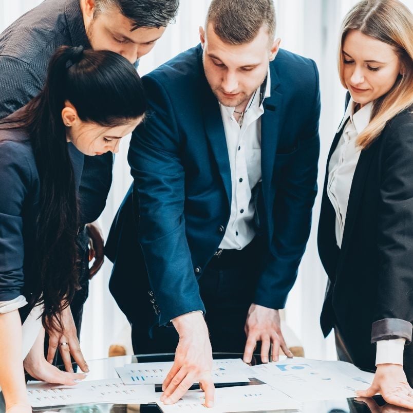 Four business professionals in formal attire are gathered around a table, closely examining documents and charts as they discuss PMP certification requirements.
