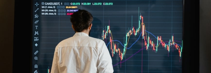 A person in a white shirt stands in front of a large screen displaying colorful cryptocurrency price charts, market data, and a release burndown chart template.