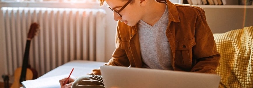 A person sits on a couch using a laptop for CAPM certification training and writing in a notebook, with a guitar leaning against a heater and books on a shelf in the background. Sunlight shines through a nearby window.