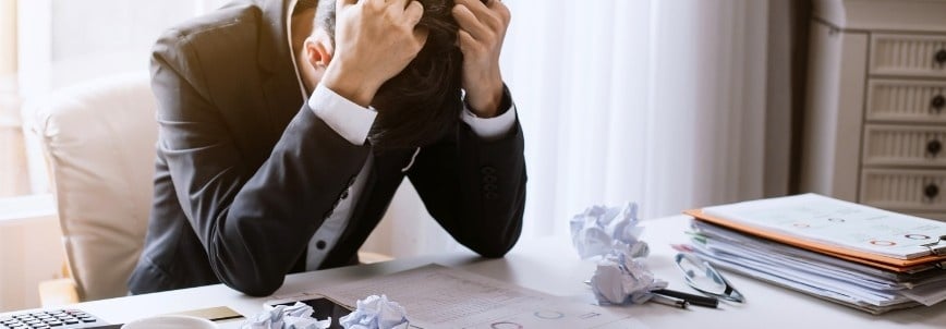 A man in a suit sits at a desk with his head in his hands, looking stressed after failing the CAPM exam. Crumpled papers, documents, and folders are scattered on the desk in front of him.