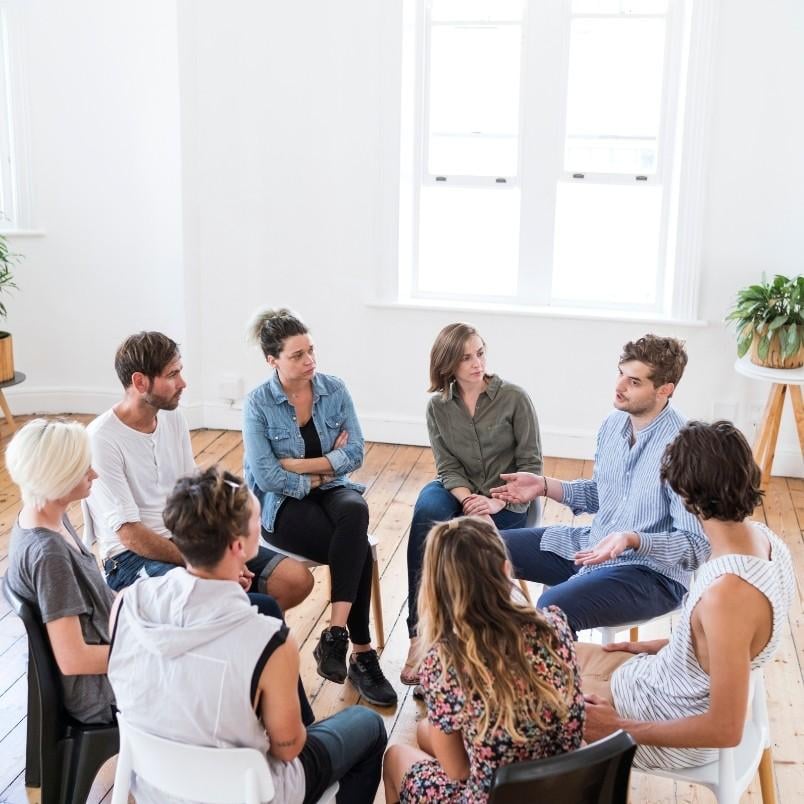 A group of eight people sits in a circle on chairs in a bright room with wooden floors, engaged in a nominal group technique discussion. Two windows and potted plants are visible in the background.