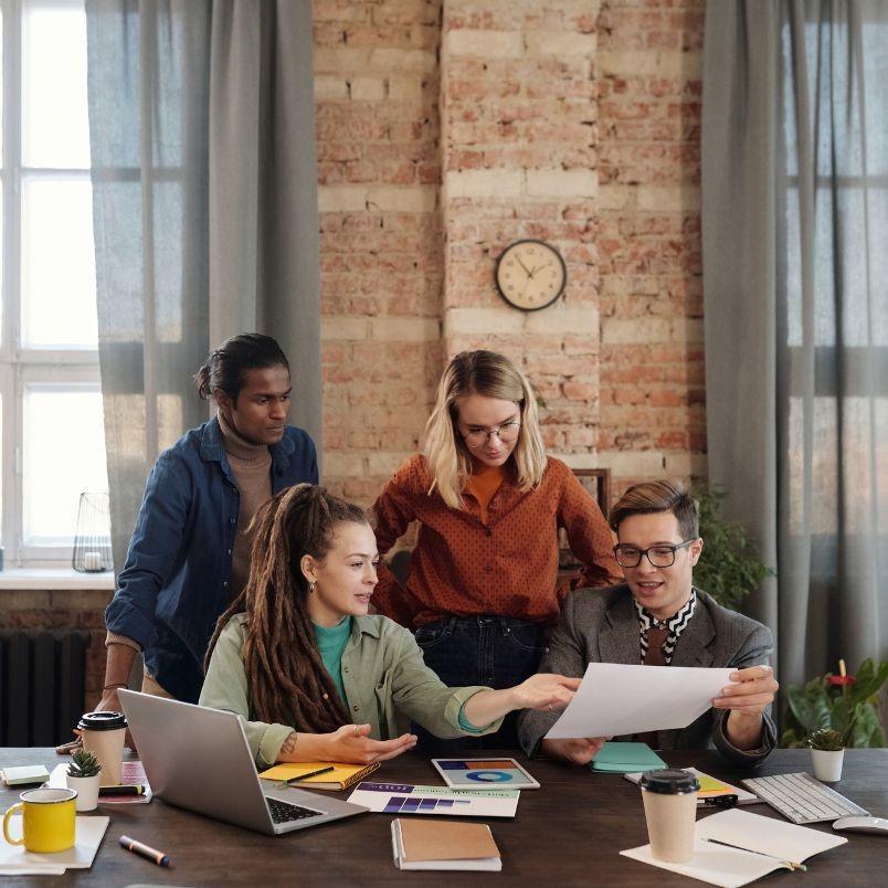 Four people gather around a table with laptops, papers, and coffee cups, discussing release instructions in a cozy office with brick walls and a clock on the wall.