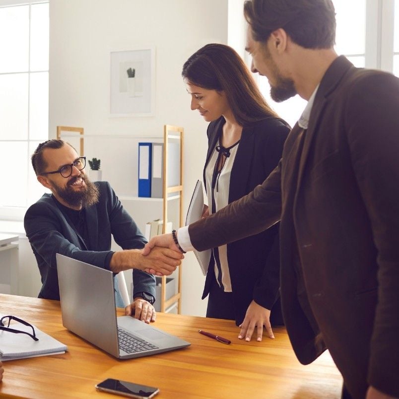 Two men and a woman in business attire greet each other in an office. One man sits at a desk with a laptop, smiling, while shaking hands with the other man, as the woman stands beside them holding documents and expert judgment instructions.