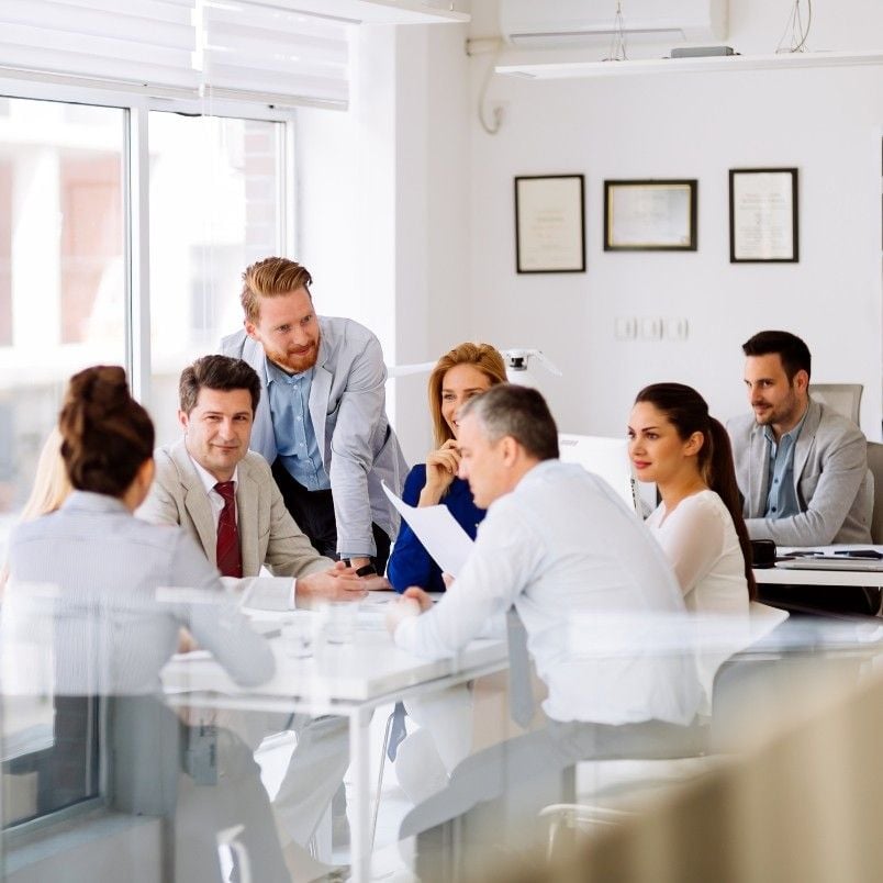 A group of eight professionals sit and stand around a conference table in a modern office, engaged in a discussion about retrospective instructions. Documents and laptops are on the table, and large windows let in natural light.