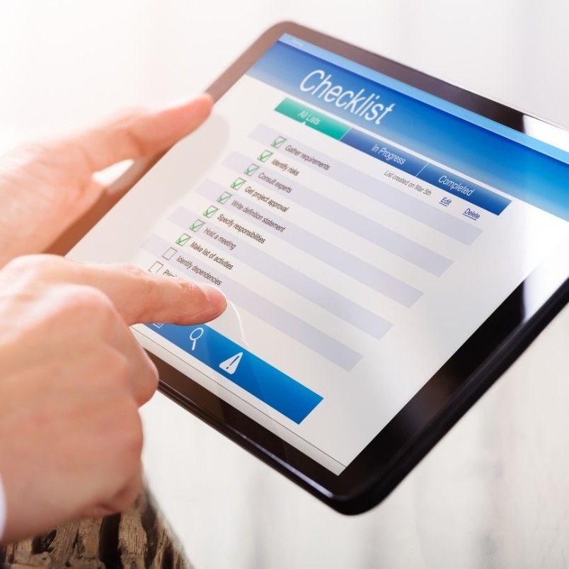 A person uses a tablet displaying a digital checklist with several tasks marked as completed, while a finger hovers over the screen to interact with the list for efficient checklist analysis.