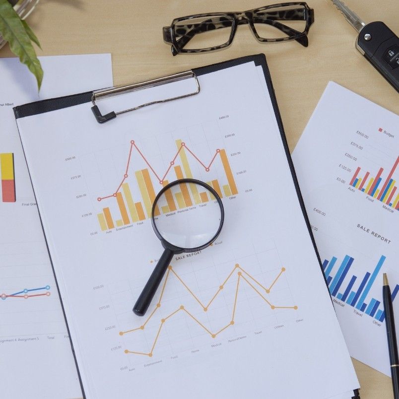 A magnifying glass rests on sales report charts with colorful graphs on a clipboard, surrounded by additional charts, eyeglasses, a pen, and a car key on a wooden desk—tools ready for Root Cause Analysis.