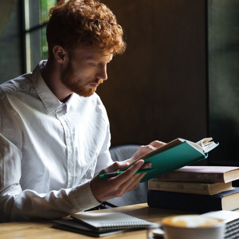 A man with curly red hair and a beard reads a green book of CAPM exam practice questions at a wooden table, with stacked books, an open notebook, and a cup of coffee nearby, in a softly lit room.