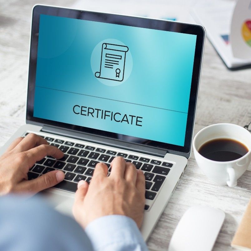 A person types on a laptop displaying an image of a certificate icon and the word "CERTIFICATE" on the screen, likely completing their CAPM certification. A cup of black coffee sits nearby on the desk.