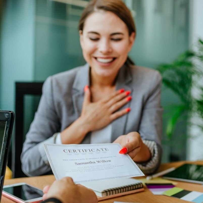 A smiling woman with bright red nails accepts a CAPM Certification Worth It certificate with her left hand while her right hand rests on her chest, sitting at a desk across from another person in a modern office setting.
