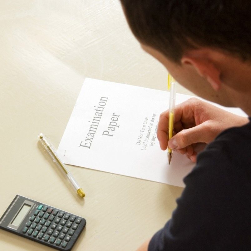 A person is holding a pencil and working through CAPM exam practice questions on an examination paper. A pen and a calculator rest on the light-colored desk beside the paper. The person's face is partially visible.