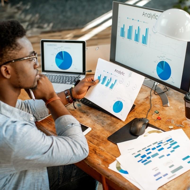 A person sits at a desk analyzing charts and graphs on multiple devices, using expert judgment to interpret blue analytics data and follow detailed instructions related to business or financial performance.