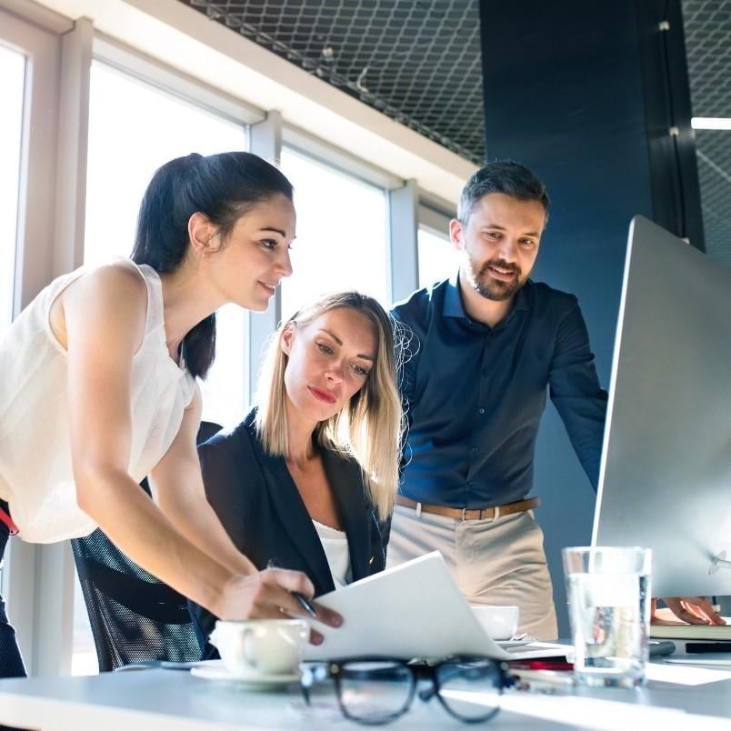 Three colleagues collaborate at a desk in a modern office, reviewing materials for CAPM certification on a computer screen. One woman points at a document, while the others watch attentively. Glasses, a cup, and water are on the table.