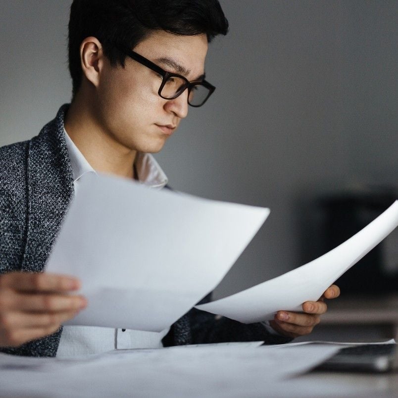 A man wearing glasses and a blazer reviews paperwork at a desk, focusing intently on CAPM job opportunities. The background is blurred, drawing attention to his concentration on the documents in his hands.