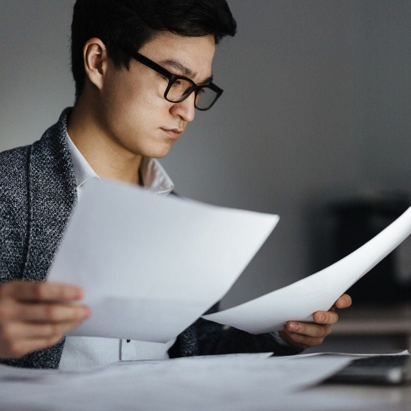 A person wearing glasses and a blazer reviews and holds several sheets of paper at a desk, appearing focused and engaged as they follow expert judgment instructions in office or academic work.