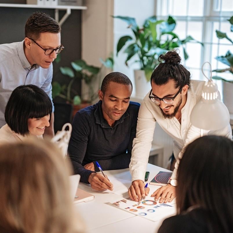 Five people gather around a table in a bright office, collaborating and smiling while reviewing colorful charts from their CAPM certification training. Green plants and a window add to the lively atmosphere in the background.