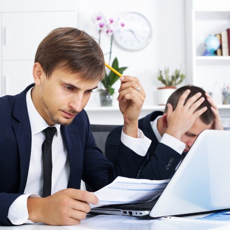 Two men in suits sit at a desk with a laptop and papers; one looks stressed while reviewing the scope statement, holding a pencil to his head, while the other has his head in his hands in frustration.
