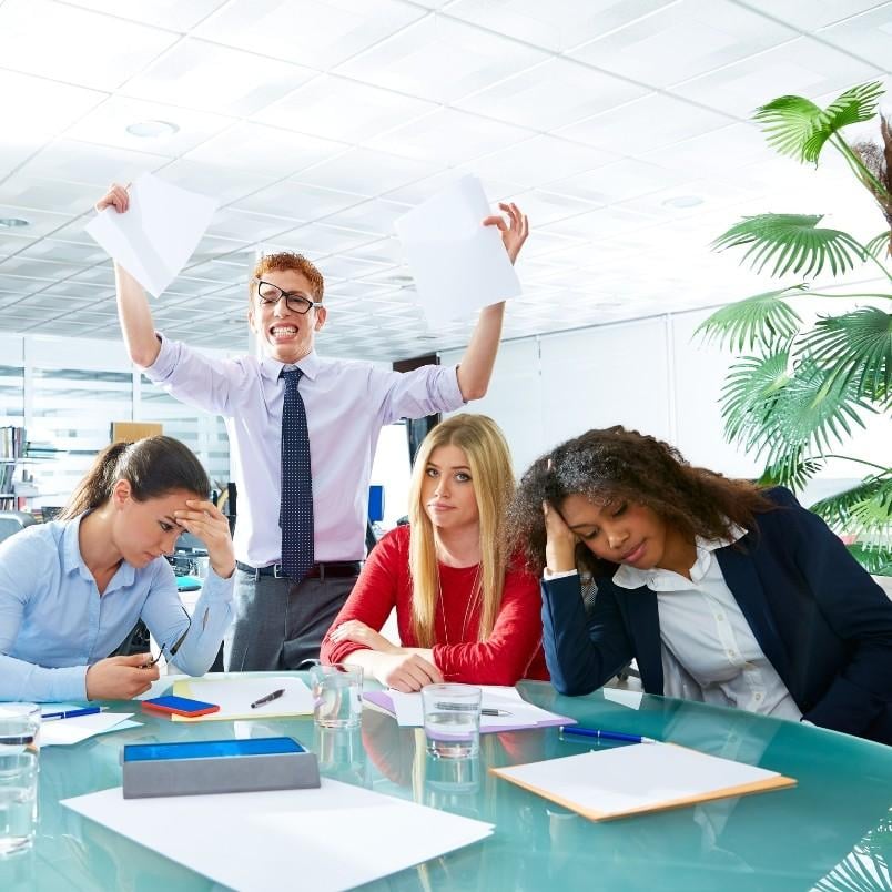 A man stands behind three women at a conference table, holding papers and smiling enthusiastically as he shares retrospective instructions. The women appear tired or stressed, with heads resting on hands in an office filled with papers and plants.
