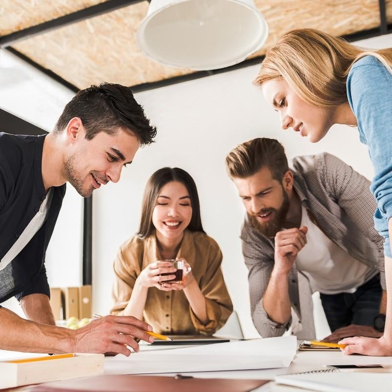 Four young adults, two men and two women, gather around a table in a modern office, smiling and discussing documents while reviewing an epic template, appearing engaged in a collaborative meeting.