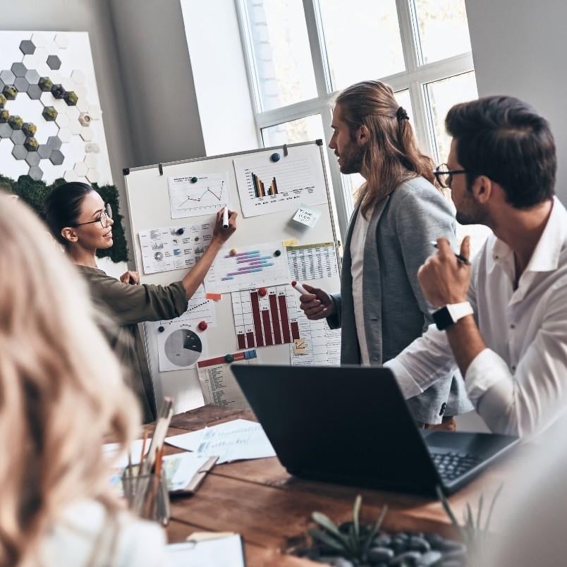 Four people in a modern office discuss data charts on a whiteboard, focusing on risk burndown chart instructions. One woman points to a graph while others listen and take notes. Sunlight streams through large windows onto the table.