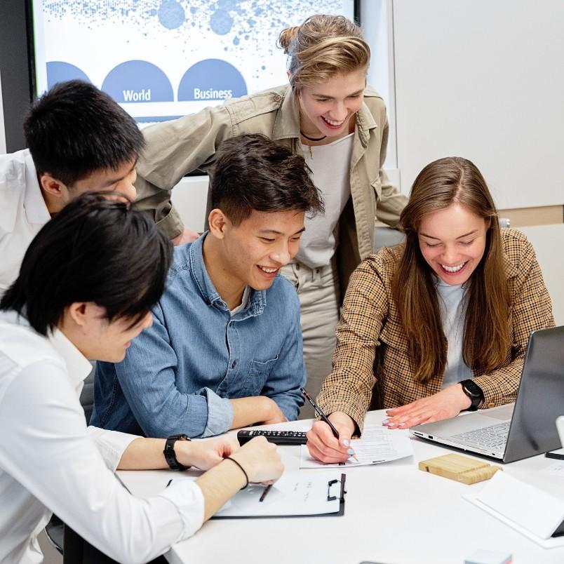Five young adults gather around a table, smiling and working together on documents and a laptop, appearing engaged and collaborative as they discuss product roadmap instructions in a modern office setting.