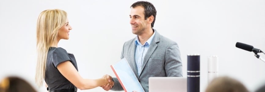 A woman and a man are shaking hands and smiling at each other in a professional setting, perhaps discussing if CAPM Certification is worth it. The man holds a folder, and office equipment with a microphone sits on the desk nearby.