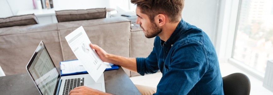 A man sits at a desk at home, reviewing a printed document with charts on capm vs other certifications while working on his laptop. A sofa and large windows are visible in the background, creating a bright workspace.
