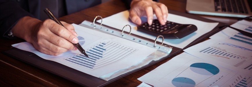 A person reviews and marks financial charts and graphs in a binder with one hand while using a calculator with the other, applying design for x principles, with a laptop nearby on the desk.
