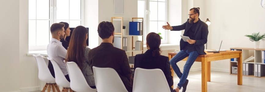 A man sits on a desk, speaking to a group of five seated adults in a bright office setting, suggesting a PROJECT COMMUNICATION meeting, lecture, or presentation.