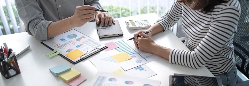 Two people sit at a desk reviewing financial documents and charts, using an epic template alongside colorful sticky notes, a tablet, a calculator, and pens—suggesting a collaborative business discussion.