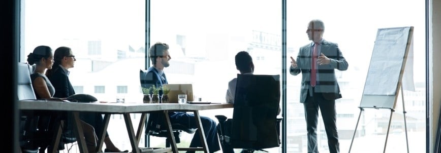 A group of four people sit at a conference table in an office while a man in a suit presents information on how to pass the CAPM exam using a flip chart, set near large windows with a city view.