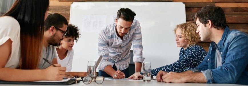 Five people sit around a table in a meeting room using the nominal group technique, discussing and reviewing documents. One person stands and writes on paper while the others listen and engage. A whiteboard is visible in the background.