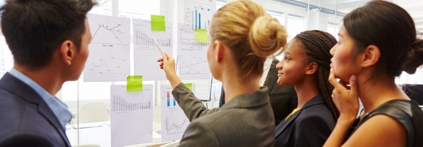Four people in business attire stand together in an office, reviewing charts and graphs from a recent ROSEMET CAPM Training. One person points at a chart as the group discusses the data and key insights on the glass wall.